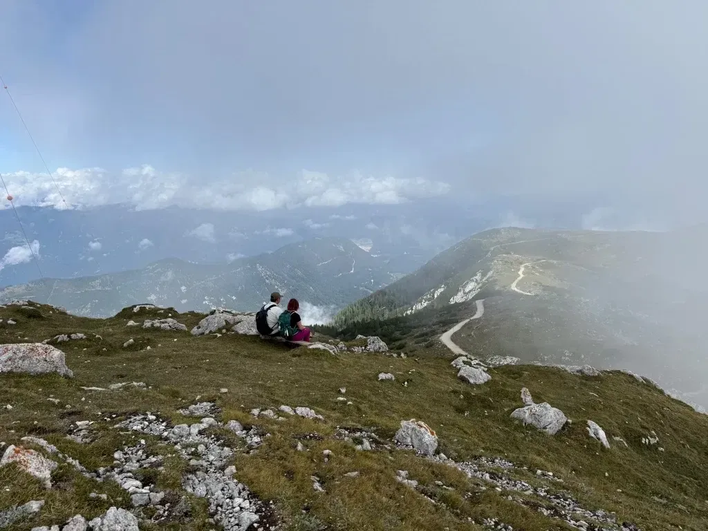 Lynn en Jeroen zitten op een rots op een grasrijke bergtop, met uitzicht over dalen, een slingerende bergweg en laaghangende wolken