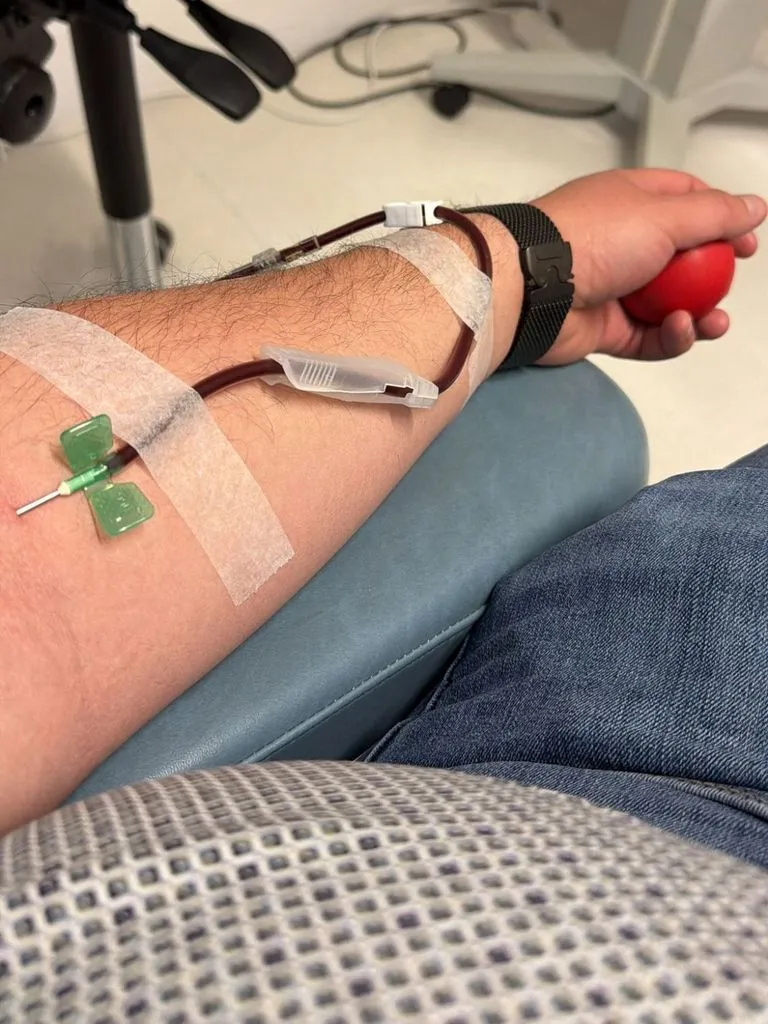 Onderarm met een donatienaald en een slangetje, vastgezet met tape, terwijl de hand een rood knijpballetje vasthoudt